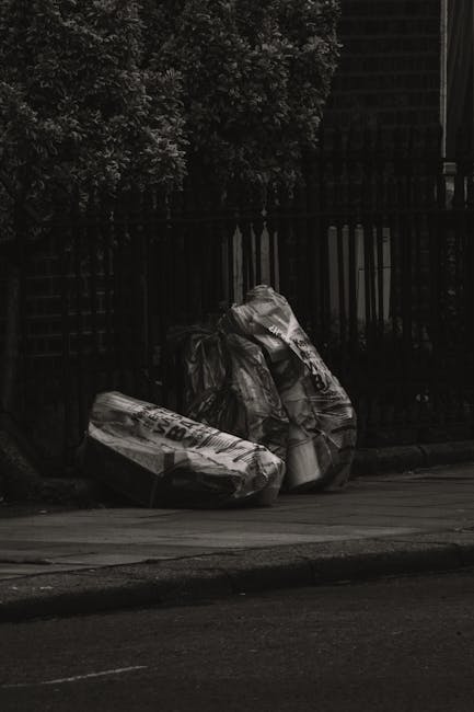 A group of large black plastic rubbish bags, filled and tied at the top, are stacked against a black metal fence on the pavement. The bags appear to contain mixed waste materials, possibly garden or h
