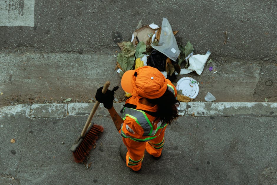 A group of large black plastic rubbish bags, filled and tied at the top, are stacked against a black metal fence on the pavement. The bags appear to contain mixed waste materials, possibly garden or h