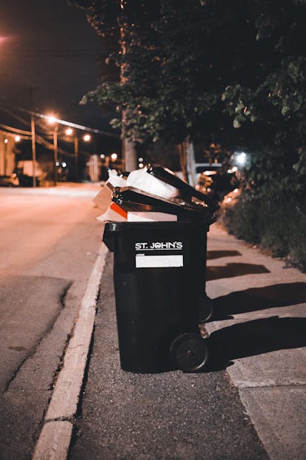 A black wheeled rubbish bin positioned on a paved sidewalk next to a curb, with the label 'St. John’s' visible on the front. The bin is partially open, revealing discarded paper and other waste mate